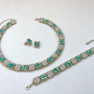 Op de afbeelding: Een set turquoise en witte kralen sieraden, inclusief een ketting, armband en oorbellen. De sieraden zijn gemaakt met vierkante kralen en zilverkleurig metaal.