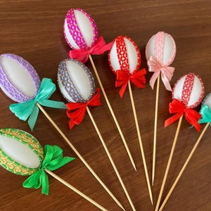Pode incluir: Sete ovos de Páscoa decorados em paus de madeira com fitas coloridas. Os ovos são brancos com diferentes padrões e cores, incluindo rosa, roxo, azul e verde.