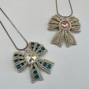 May include: Two silver beaded bow necklaces with crystal accents. One bow is teal and the other is pink.