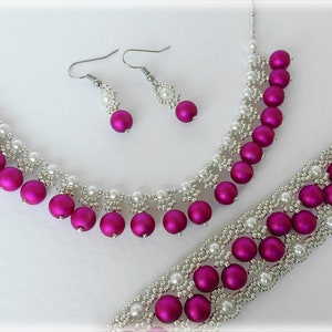 Puede incluir: Un conjunto de joyas de cuentas rosa y blancas con un collar, una pulsera y pendientes. El collar y la pulsera presentan una delicada cadena plateada con pequeñas cuentas blancas y cuentas rosas más grandes. Los pendientes son plateados con una sola cuenta rosa.