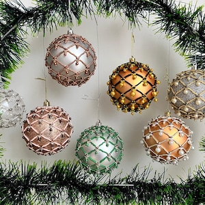 Könnte beinhalten: Sieben Weihnachtskugeln mit aufwendigen Perlenmustern in verschiedenen Farben, darunter Silber, Gold, Bronze und Grün. Sie hängen vor einem Hintergrund aus grüner Girlande. Jede Kugel hat ein einzigartiges Muster.