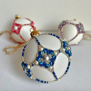 May include: Three white Christmas baubles with beaded designs. The bauble on the left has pink beads, the bauble in the centre has blue and white beads, and the bauble on the right has purple beads.