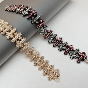 May include: Two beaded bracelets. One is gold-toned, the other is black, red, and silver. Both bracelets feature a repeating cross-like pattern. The gold bracelet has a small gold-toned clasp. The other bracelet has a silver clasp.