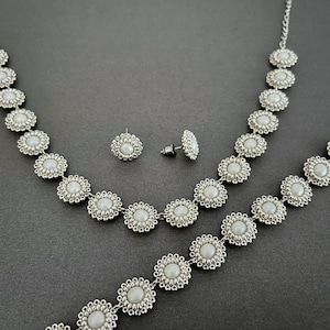 May include: A silver necklace, bracelet, and earrings set with white pearl-like beads. The jewelry features a circular design with a central pearl-like bead.