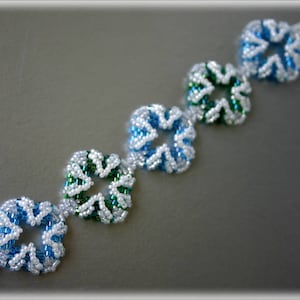 Könnte beinhalten: Ein weißes und blaues Perlenarmband mit grünen Akzenten. Das Armband besteht aus kleinen, quadratischen Perlen, die in einem sich wiederholenden Muster angeordnet sind.