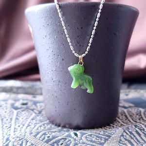 May include: A green jade pendant shaped like a small animal, hanging from a silver chain. The pendant is on a grey ceramic surface.