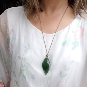 May include: A green jade leaf pendant necklace on a brown cord. The pendant is carved in the shape of a leaf with intricate details.