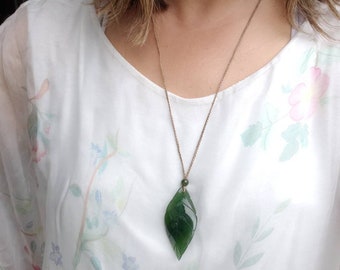 May include: A green jade leaf pendant necklace on a brown cord. The pendant is carved in the shape of a leaf with intricate details.