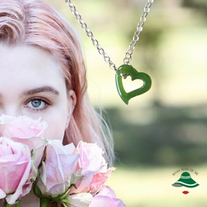 May include: A close-up of a woman's face with pink hair, partially obscured by a bouquet of pink roses. A silver chain necklace with a green jade heart pendant hangs in the background.