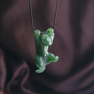 Può includere: Un ciondolo di giada verde a forma di gatto, appeso a un cordino nero. Il gatto è scolpito con dettagli lisci e la giada ha una qualità traslucida. Il ciondolo è sospeso su uno sfondo scuro e sfocato.