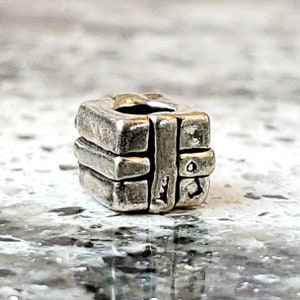 May include: A silver charm in the shape of a small gift box with a ribbon. The charm is detailed with textured lines and a small hole for attaching to a bracelet.