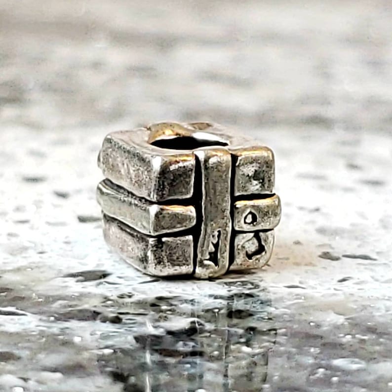 May include: A silver charm in the shape of a small gift box with a ribbon. The charm is detailed with textured lines and a small hole for attaching to a bracelet.