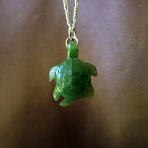 May include: A green jade turtle pendant on a gold chain. The turtle is carved in detail and has a smooth, polished finish.