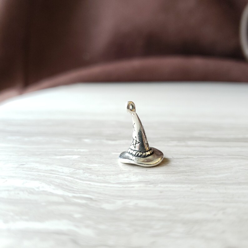 May include: A silver charm in the shape of a witch's hat. The hat has a pointed top and a wide brim. The charm is detailed with a star pattern and a rope-like band around the brim.