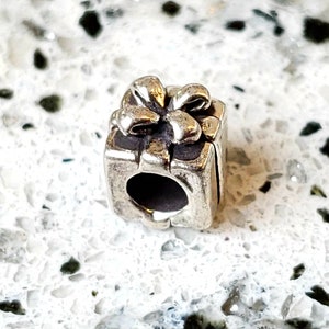 May include: Silver charm in the shape of a gift box with a bow on top. The charm has a hole in the side for attaching to a bracelet.