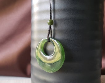 May include: A green jade pendant necklace with a round, open circle design. The pendant is hanging from a black cord with a green bead.