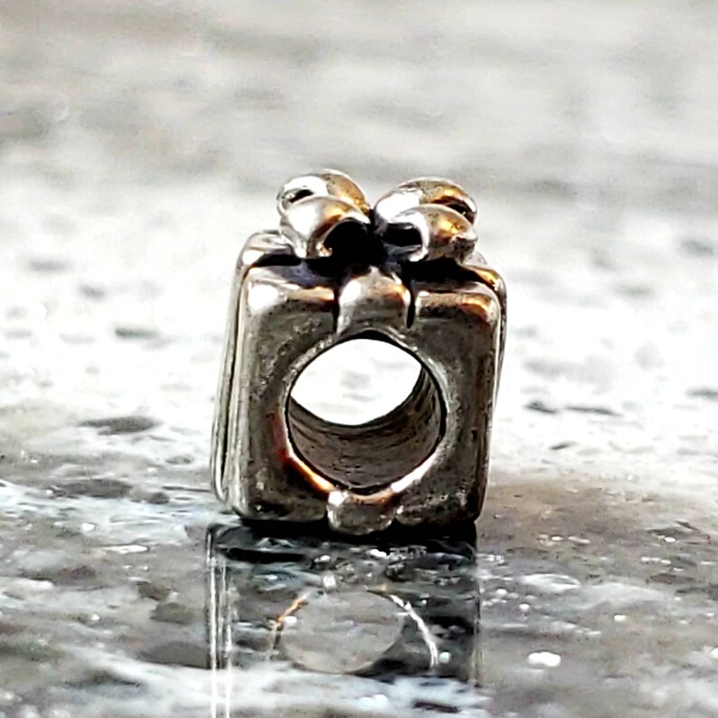 May include: Silver charm in the shape of a gift box with a bow on top. The charm has a hole in the center for attaching to a bracelet.