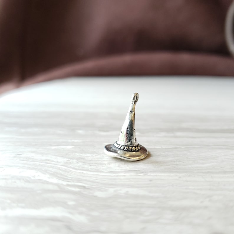 May include: A silver charm in the shape of a witch's hat. The hat has a pointed top and a wide brim. The charm is detailed with a star pattern on the hat.