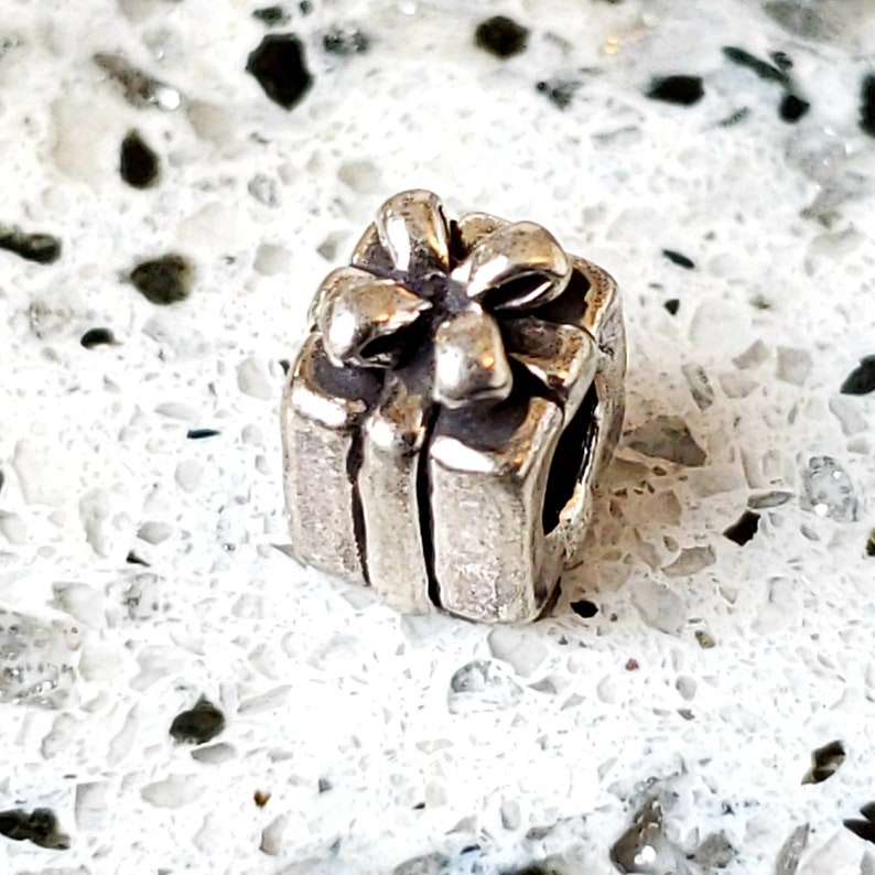 May include: A silver charm in the shape of a gift box with a bow on top. The charm is designed to be used on a bracelet or necklace.