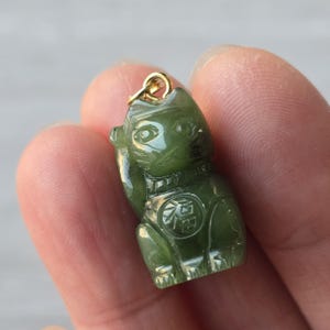 Puede incluir: Un dije de Maneki Neko de jade verde con una fianza dorada. El dije está tallado en forma de gato sentado con una pata levantada. Un emblema circular con caracteres chinos está en el pecho del gato.