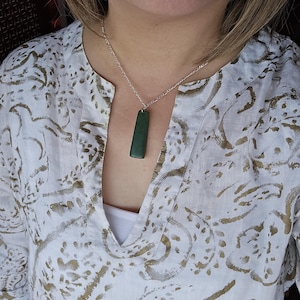 May include: A dark green, rectangular stone pendant hangs from a silver-coloured metal chain. The pendant has a smooth, matte finish and is suspended by a small metal bail. The necklace is worn over a white shirt featuring a gold-toned leaf pattern.