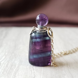 Könnte beinhalten: Eine silberne Kettenkette mit einem Anhänger in Form einer violetten Fluoritflasche. Die Flasche hat einen runden, violetten Amethystverschluss.