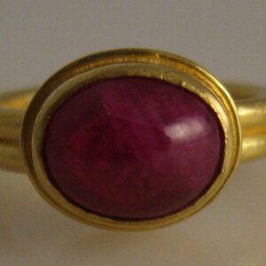 May include: A gold ring featuring a large, oval-shaped, deep red gemstone. The gemstone is set in a gold bezel, and the ring band has a double-band design. The ring is a piece of jewelry.