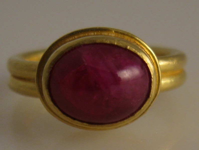 May include: A gold ring featuring a large, oval-shaped, deep red gemstone. The gemstone is set in a gold bezel, and the ring band has a double-band design. The ring is a piece of jewelry.