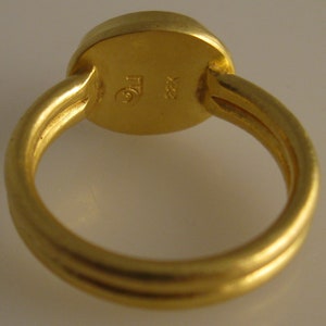 May include: A close-up of a gold ring with a round, flat top. The ring band has a triple-band design. The ring is stamped with the markings "TG" and "22K".