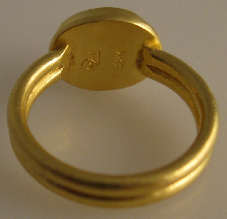 May include: A close-up of a gold ring with a round, flat top. The ring band has a triple-band design. The ring is stamped with the markings "TG" and "22K".