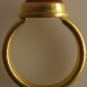 May include: A gold ring with a large, round, red gemstone. The ring's band is a simple, polished gold, and the gemstone is set in a gold bezel. The ring is a classic design, suitable for various occasions.