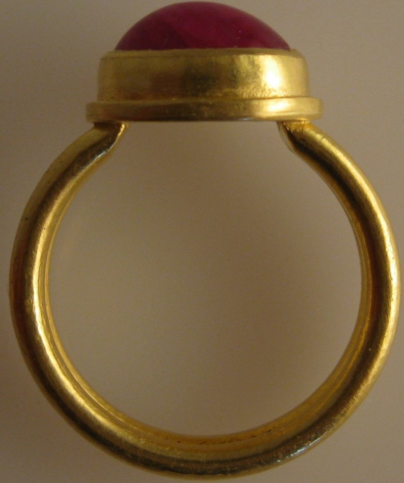 May include: A gold ring with a large, round, red gemstone. The ring's band is a simple, polished gold, and the gemstone is set in a gold bezel. The ring is a classic design, suitable for various occasions.
