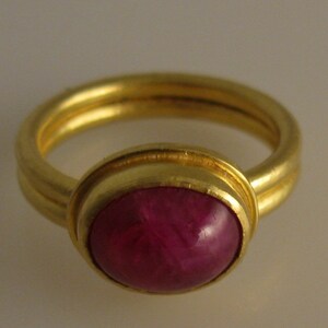 May include: A gold ring featuring a large, oval-shaped, deep red gemstone. The ring has a double band design and a polished finish, showcasing the gemstone's rich color and luster. A classic piece of jewelry.