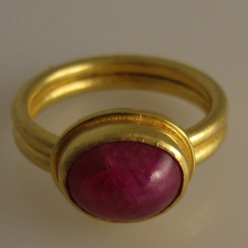 May include: A gold ring featuring a large, oval-shaped, deep red gemstone. The ring has a double band design and a polished finish, showcasing the gemstone's rich color and luster. A classic piece of jewelry.