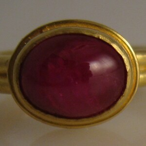 May include: A gold ring featuring a large, oval-shaped, deep red gemstone. The gemstone is set in a gold bezel, and the ring band has a double-band design. The ring's design is simple and elegant, suitable for everyday wear or special occasions.