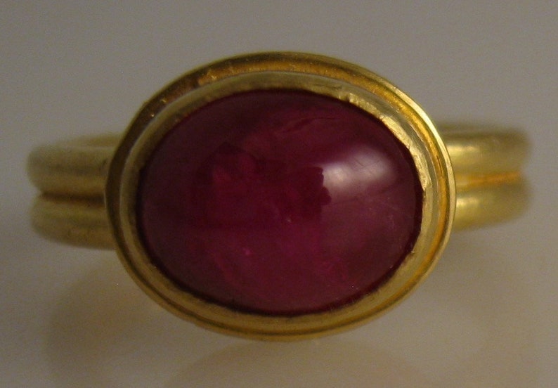 May include: A gold ring featuring a large, oval-shaped, deep red gemstone. The gemstone is set in a gold bezel, and the ring band has a double-band design. The ring's design is simple and elegant, suitable for everyday wear or special occasions.