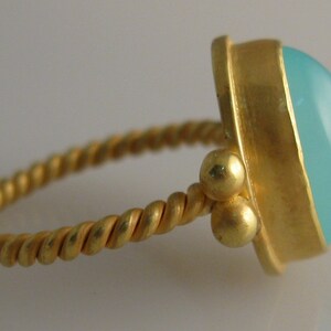 May include: A gold ring with a twisted band and a light blue gemstone. The ring has three small gold beads on the side.