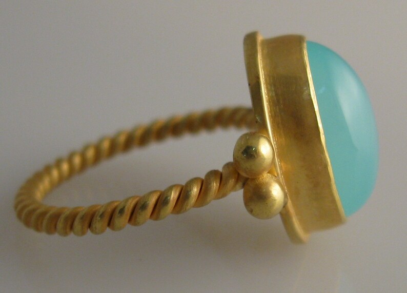 May include: A gold ring with a twisted band and a light blue gemstone. The ring has three small gold beads on the side.