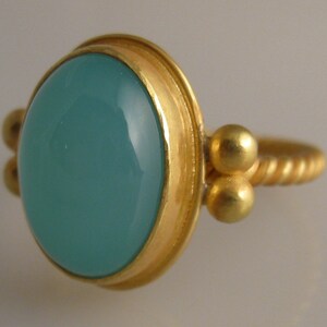 May include: A gold ring with a light blue oval gemstone set in a bezel setting. The band is twisted and has two small gold balls on each side of the stone.