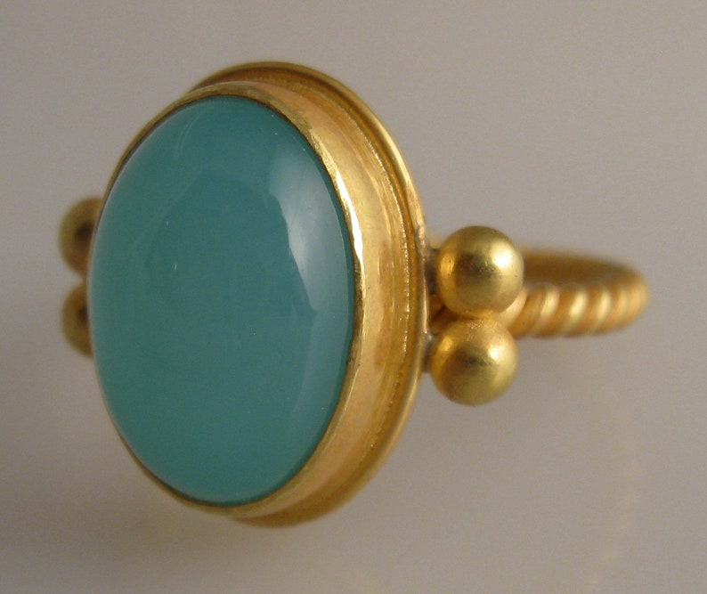 May include: A gold ring with a light blue oval gemstone set in a bezel setting. The band is twisted and has two small gold balls on each side of the stone.