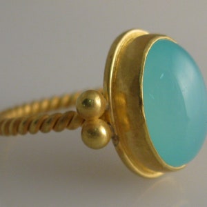 May include: A gold ring with a twisted band and a light blue gemstone set in a gold bezel. The ring has two small gold balls on the band.