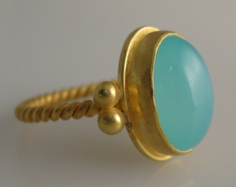 May include: A gold ring with a twisted band and a light blue gemstone set in a gold bezel. The ring has two small gold balls on the band.