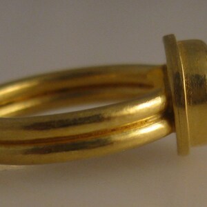 May include: A gold ring with a double band and a large, round, red gemstone. The gemstone is set in a square bezel. The ring is shown from a side view.