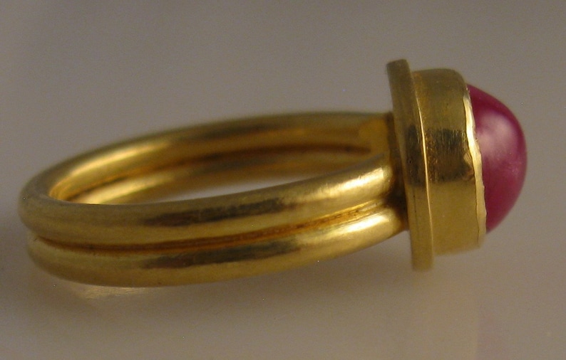 May include: A gold ring with a double band and a large, round, red gemstone. The gemstone is set in a square bezel. The ring is shown from a side view.