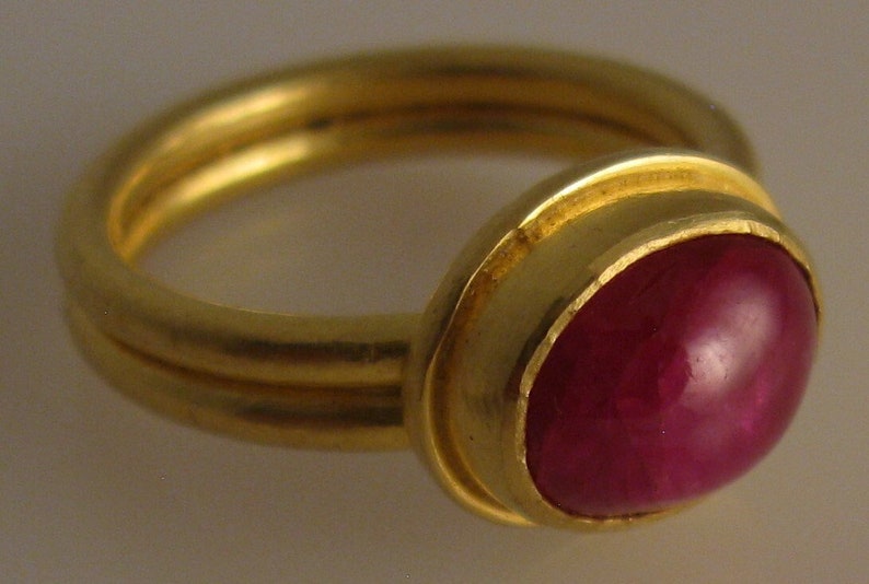 May include: A gold ring with a double band and a large, oval, deep red gemstone. The gemstone is set in a gold bezel. The ring has a classic, elegant design, suitable for various occasions. The ring is a piece of jewelry.