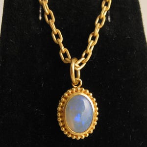 May include: A gold-colored necklace with a pendant featuring an oval opal gemstone. The opal has a blue and white color. The pendant is surrounded by a beaded gold border and hangs from a gold chain.