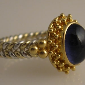 May include: A gold and silver ring with a dark blue gemstone set in a gold bezel. The ring band is a braided design with alternating gold and silver.