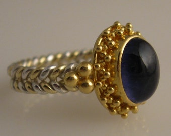 May include: A gold and silver ring with a dark blue gemstone set in a gold bezel. The ring band is a braided design with alternating gold and silver.
