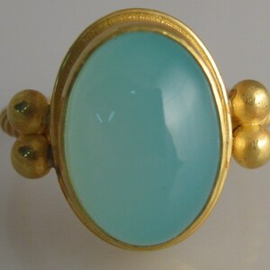 May include: A gold ring with a light blue oval gemstone set in a bezel setting. The ring has three gold beads on each side of the gemstone.