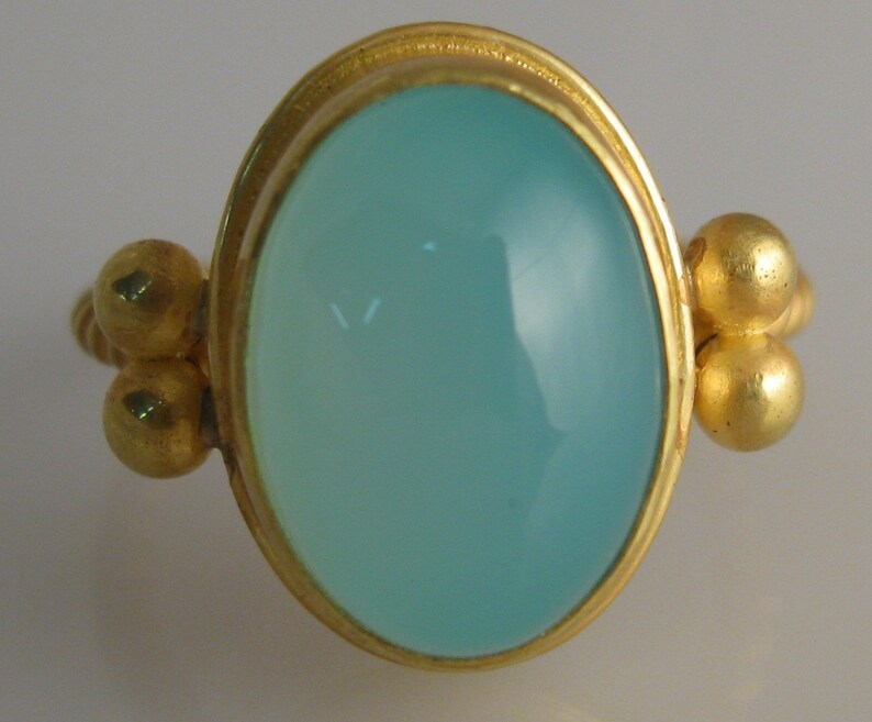 May include: A gold ring with a light blue oval gemstone set in a bezel setting. The ring has three gold beads on each side of the gemstone.
